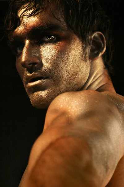 Dramatic portrait of intense looking shirtless male model in bronze and gold makeup turning