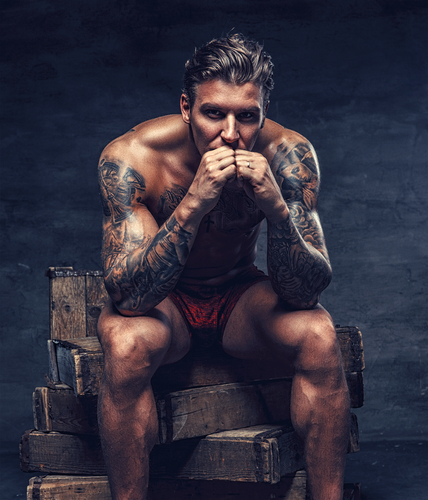 Naked muscular tattooed guy sitting on wooden boxes.